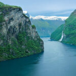 Fjord in Norwegen mit steilen, bewaldeten Ufern und ruhigem, klarem Wasser unter einem blauen Himmel.