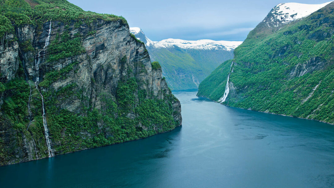 Fjord in Norwegen mit steilen, bewaldeten Ufern und ruhigem, klarem Wasser unter einem blauen Himmel.