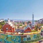 Blick von oben auf den Park Güell in Barcelona, mit bunten Mosaiken und der Stadt im Hintergrund.