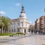 Stadtplatz mit Palmen und einem hohen Uhrturm im Hintergrund.