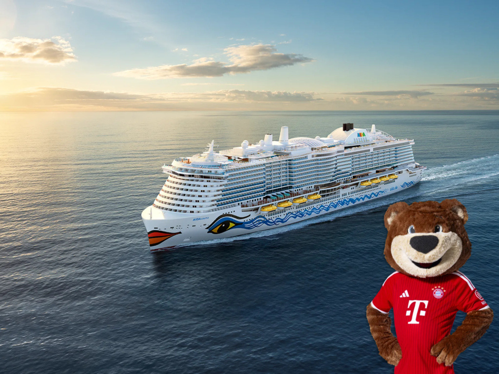 Kreuzfahrtschiff von AIDA Cruises auf dem Meer bei Sonnenuntergang, im Vordergrund rechts ein Plüschmaskottchen im roten FC-Bayern-Trikot.